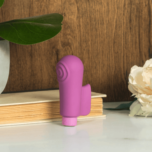 A small pink vibrator with a curved shape, standing upright on a white surface in front of stacked books, with a wooden wall background, a green leaf at left, and a cream flower at right.
