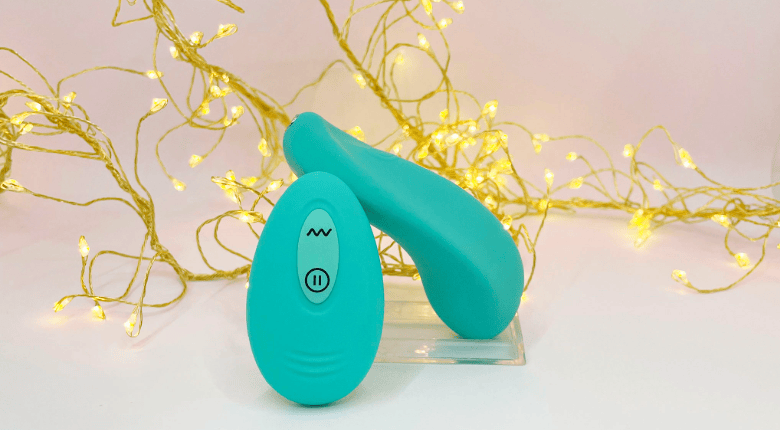 A teal silicone personal massager and its matching remote control with a power button, displayed on a clear acrylic stand against a light pink background with decorative warm white fairy lights.
