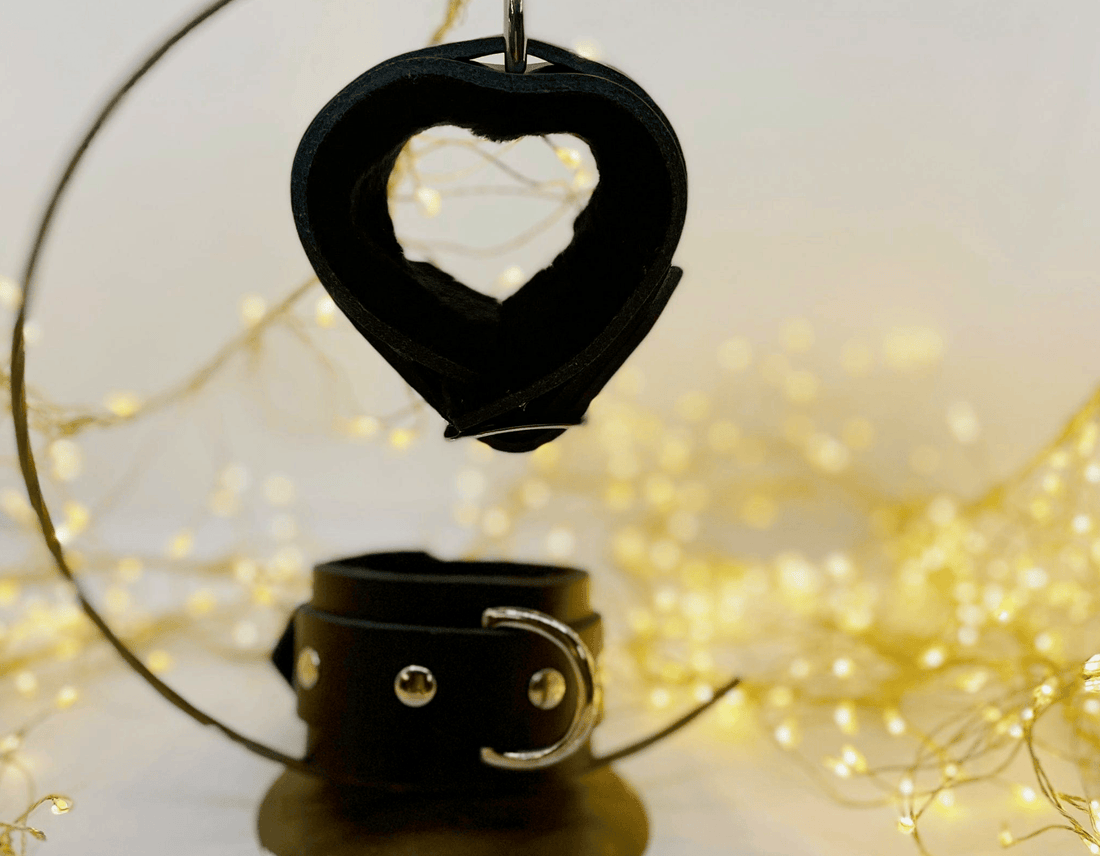 Black leather wrist restraint cuff displayed above a matching black leather cuff with silver-toned studs, set against a warm background with blurred fairy lights.