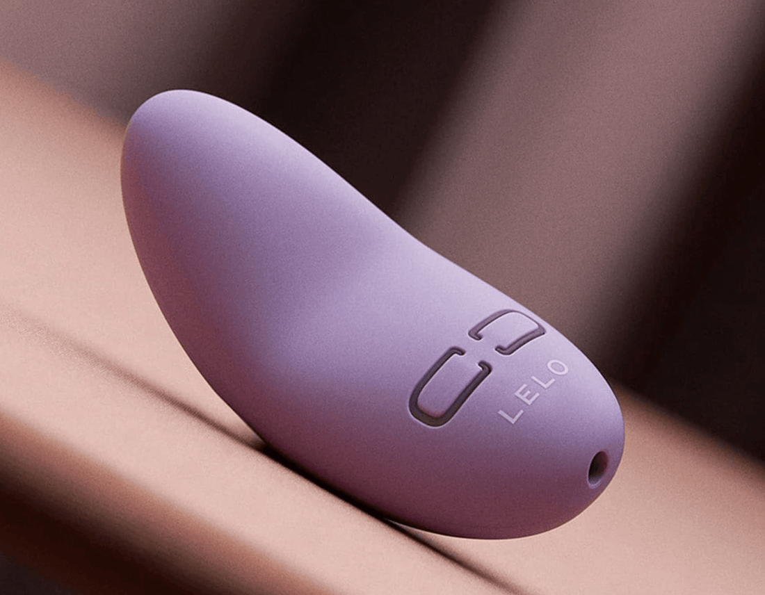 A close-up of a lavender LELO Lily silicone personal massager resting on a smooth, warm-toned surface with a blurred background.