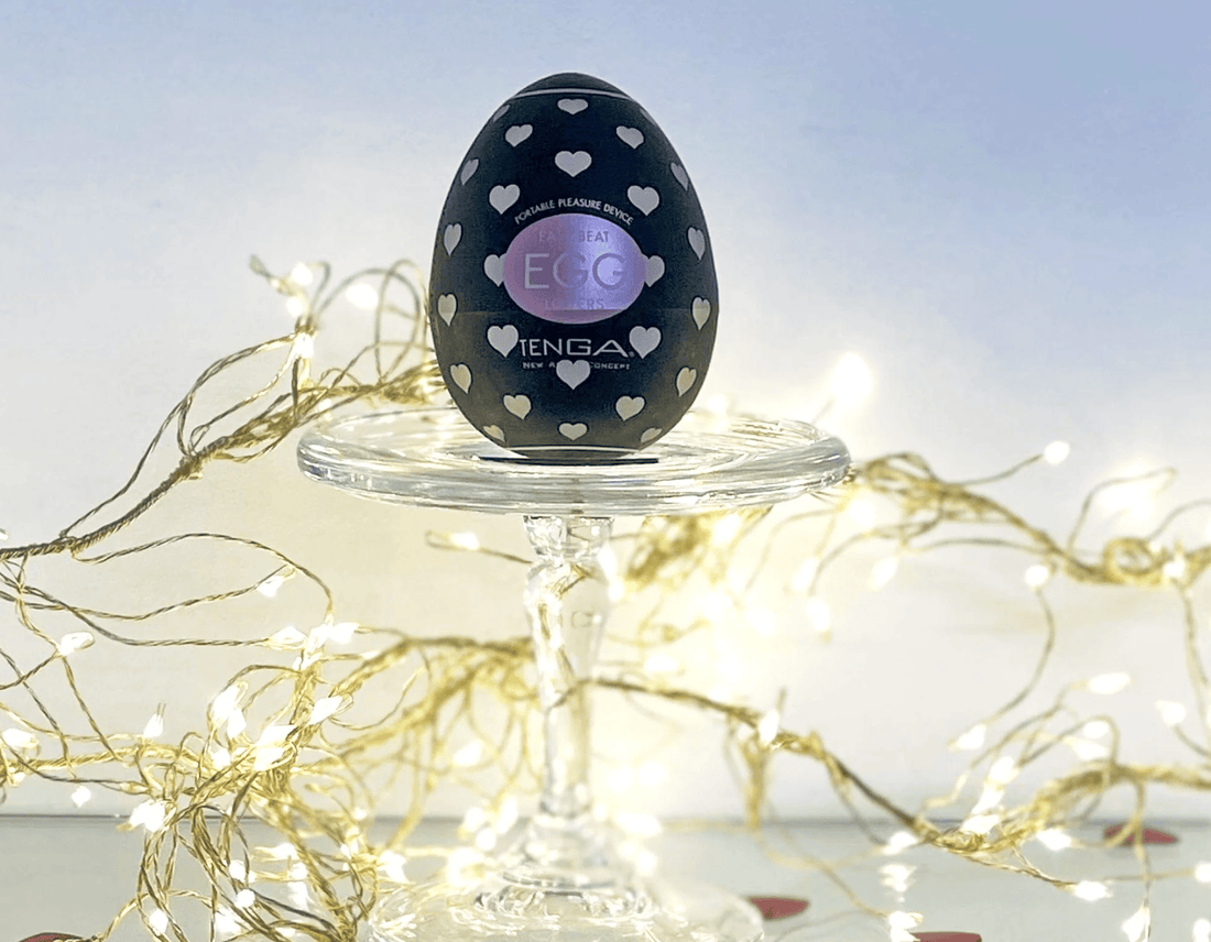 A black Tenga egg toy with white heart patterns and a purple circular label, displayed on a clear glass pedestal stand with warm fairy lights in the background.