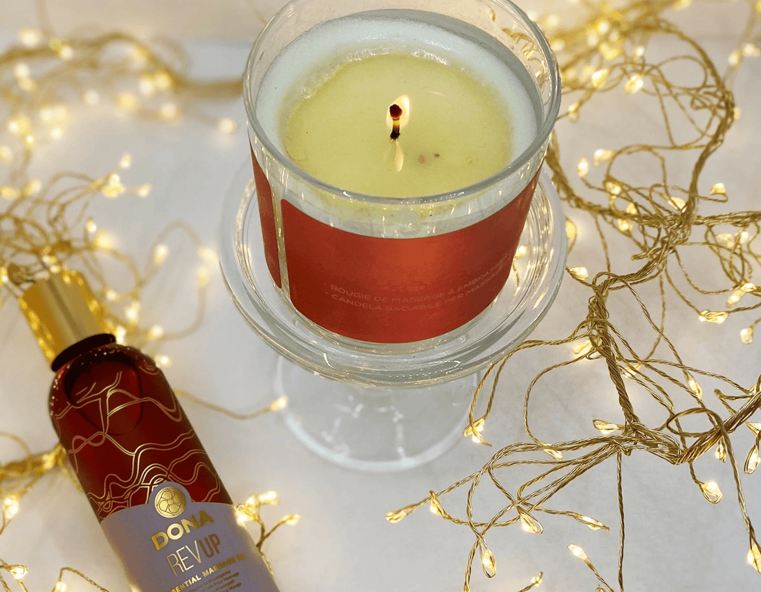 A lit massage candle in a clear glass holder with a red label sits on a glass stand, surrounded by warm fairy lights. A matching red and gold bottle of massage oil labeled 'DONA REVAIR' is placed in the lower left on a white surface.