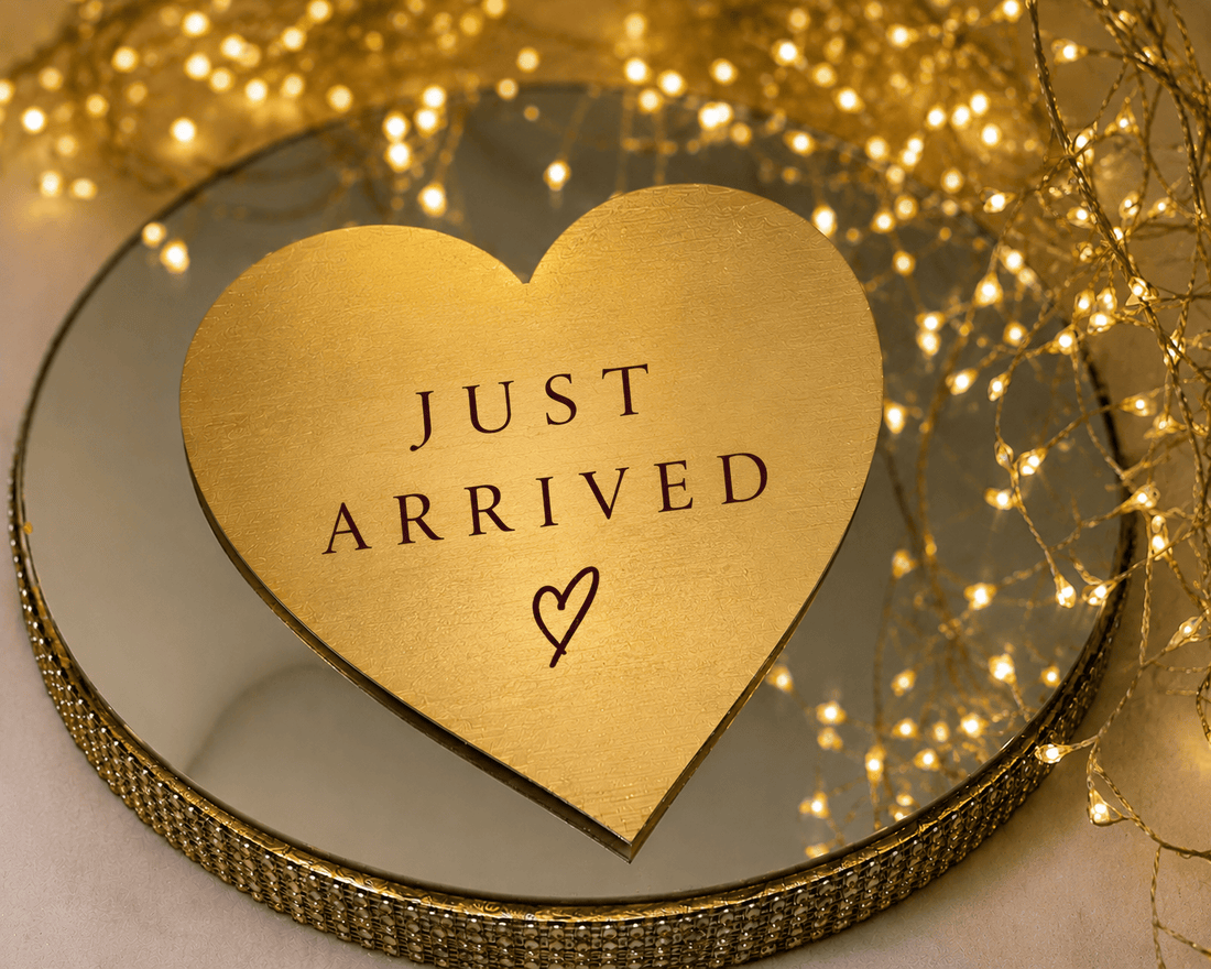 A heart-shaped gold plaque with the words 'JUST ARRIVED' and a small heart, displayed on a round mirrored tray with warm string lights in the background.
