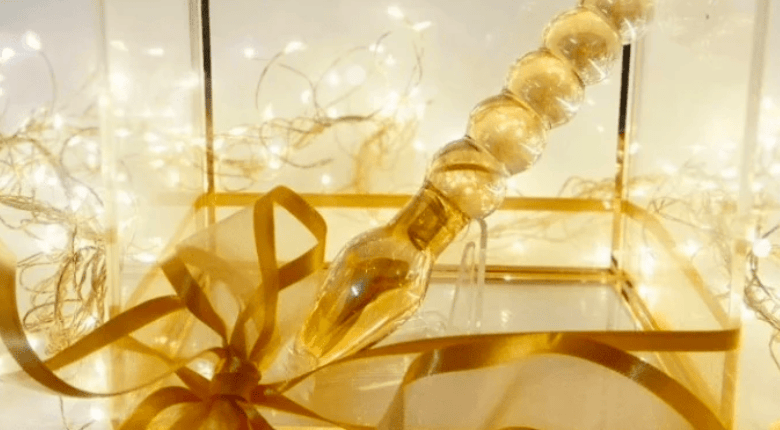 A clear acrylic gift box decorated with a gold ribbon and bow, illuminated by warm fairy lights, with a gold beaded glass dildo inside.