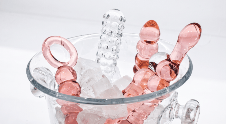 Pink and clear glass adildos arranged inside a clear glass bucket filled with ice on a white background.