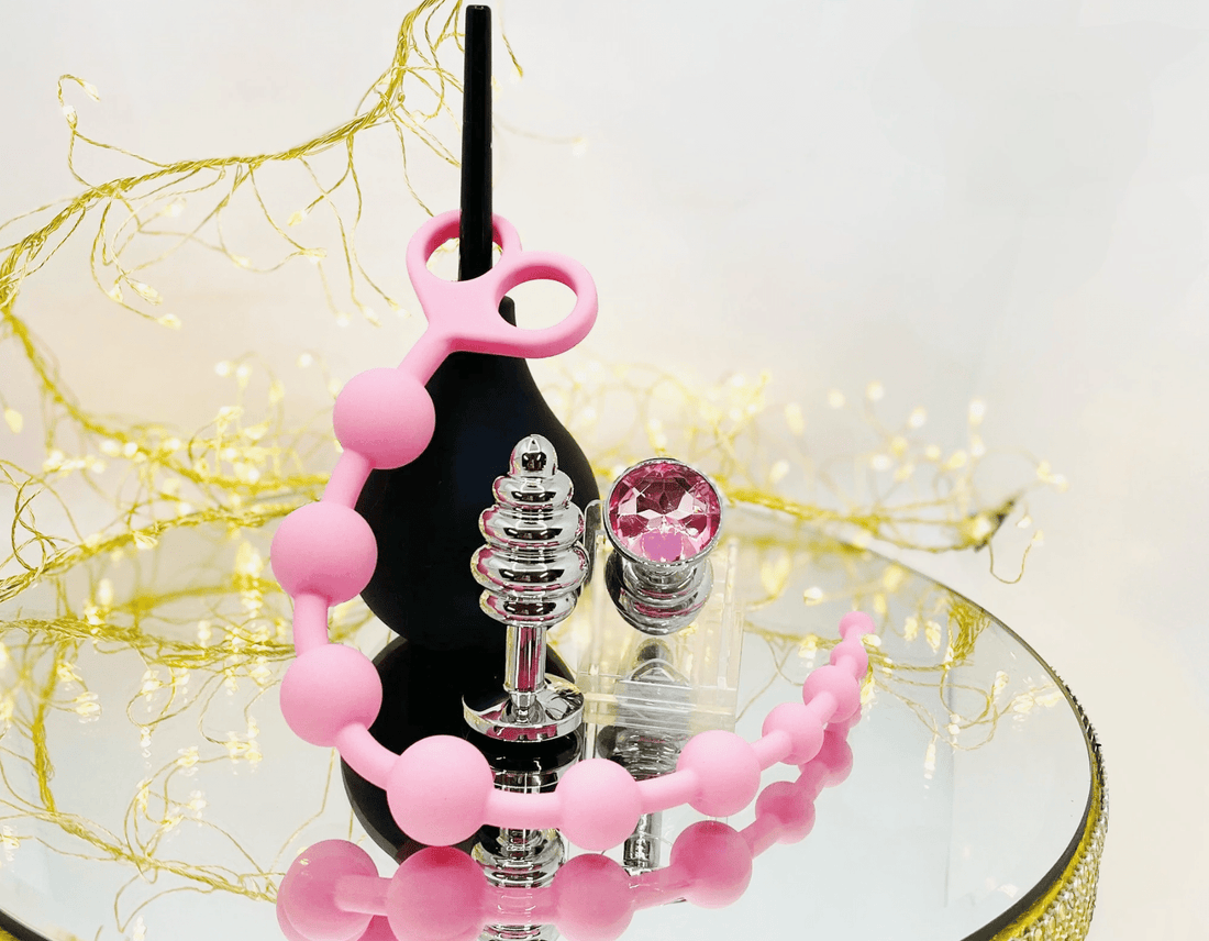 A selection of anal sex toys; Pink silicone beaded anal beads; black douche;  silver & jewelled butt plugs displayed on a mirror against a soft white background with gold fairy lights