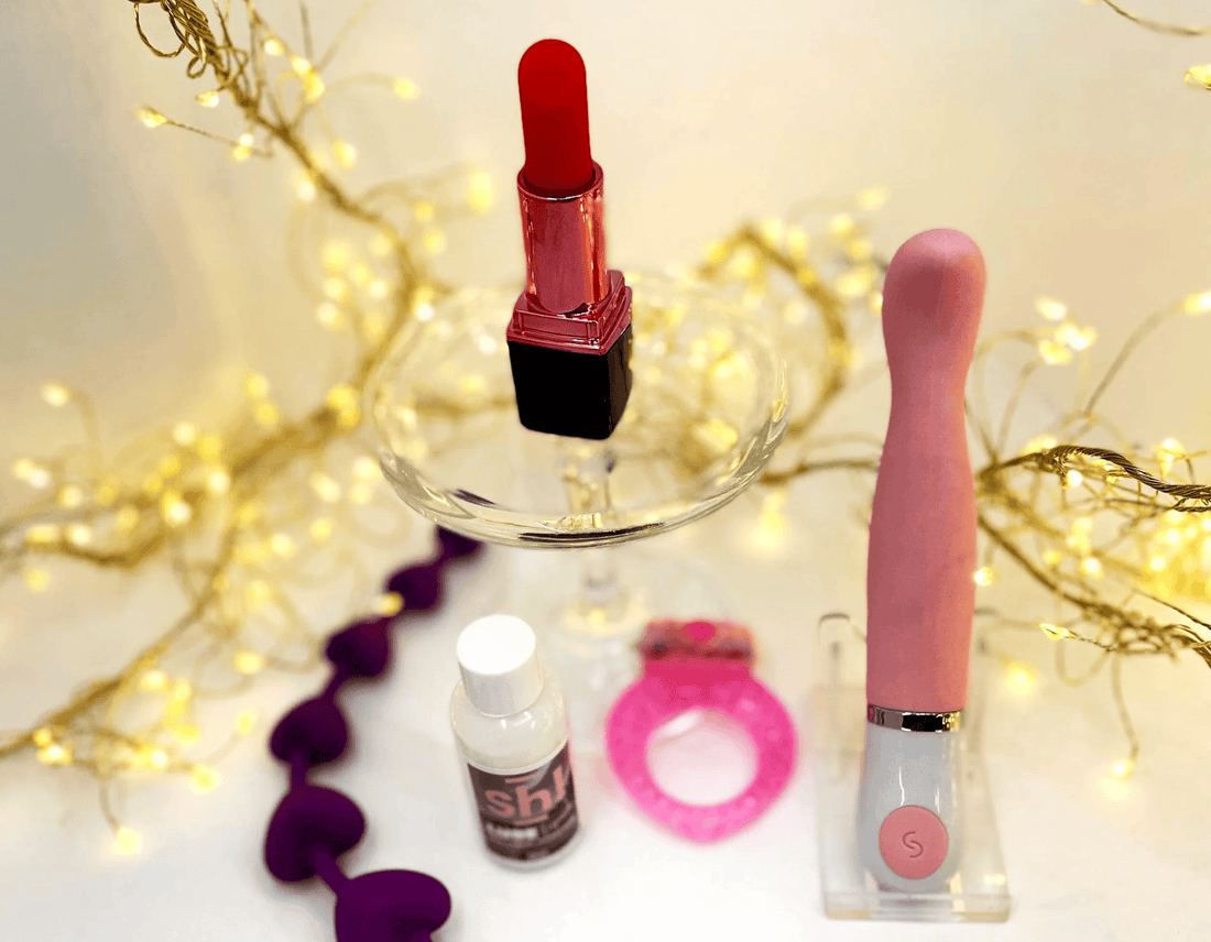 A pink personal massager in a clear acrylic stand, a red lipstick vibrator on a glass pedestal, a small bottle of Sh! Lue a pink cock-ring ring, and purple heart-shaped anal beads, styled with warm fairy lights.