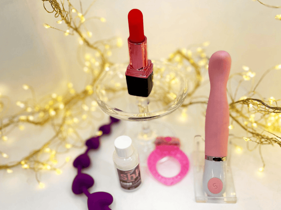 A pink g-spot vibrator with a clear acrylic stand, displayed alongside a red lipstick vibrator on a glass pedestal, a small bottle of lube, a pink cock-ring, and purple heart-shaped anal beads against a warm background with gold fairy lights.