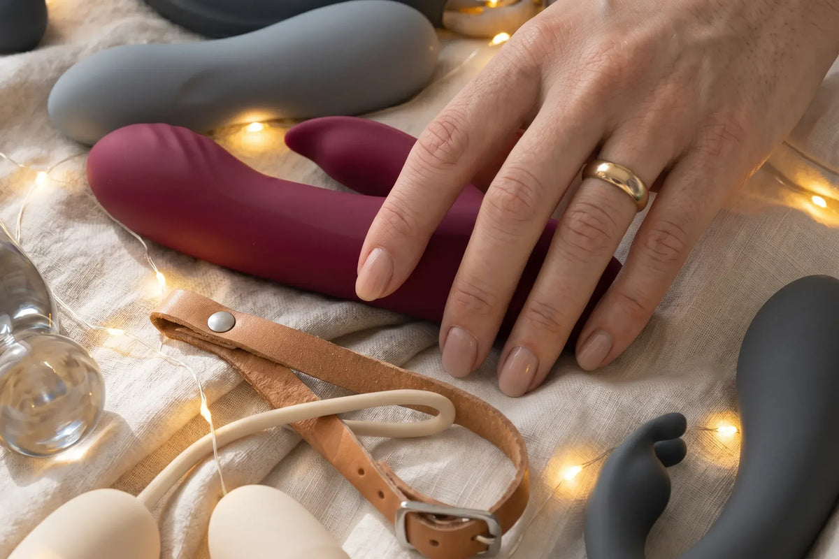 A close-up of a hand resting on a dark red silicone personal massager, surrounded by other intimate devices in gray, beige, and brown, with warm fairy lights on a textured fabric background.