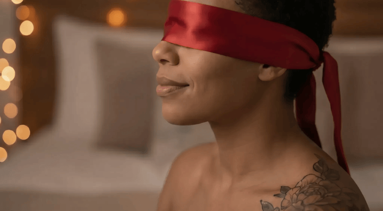 A close-up side profile of a person wearing a satin red blindfold, with warm fairy string lights and a soft neutral background.