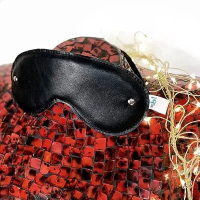 A black leather blindfold with silver metal eyelets, displayed on top of a red mosaic-patterned body with decorative fairy lights.