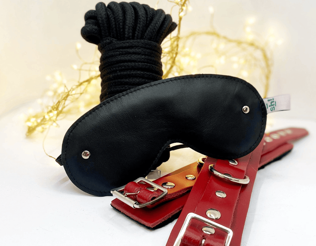 A black leather blindfold with silver metal rivets and black bondage, displayed with red leather  wrist cuffs on a white surface with warm string lights in the background.