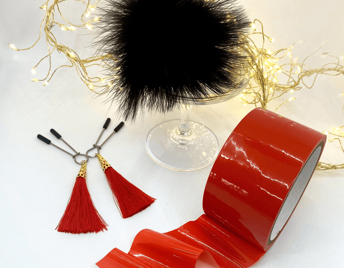 A roll of glossy red bondage tape alongside two red nipple tassels, arranged on a white surface with decorative fairy lights and a black feather pom-pom in the background.