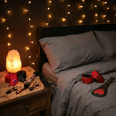 A bedroom scene with a bed made up in gray bedding, wawit red ankle cuffs & black & red heart-shaped spanker. Fairy lights hanging on the wall, and a bedside table holding a glowing salt lamp, red & black sating ties, and a leopard print whip