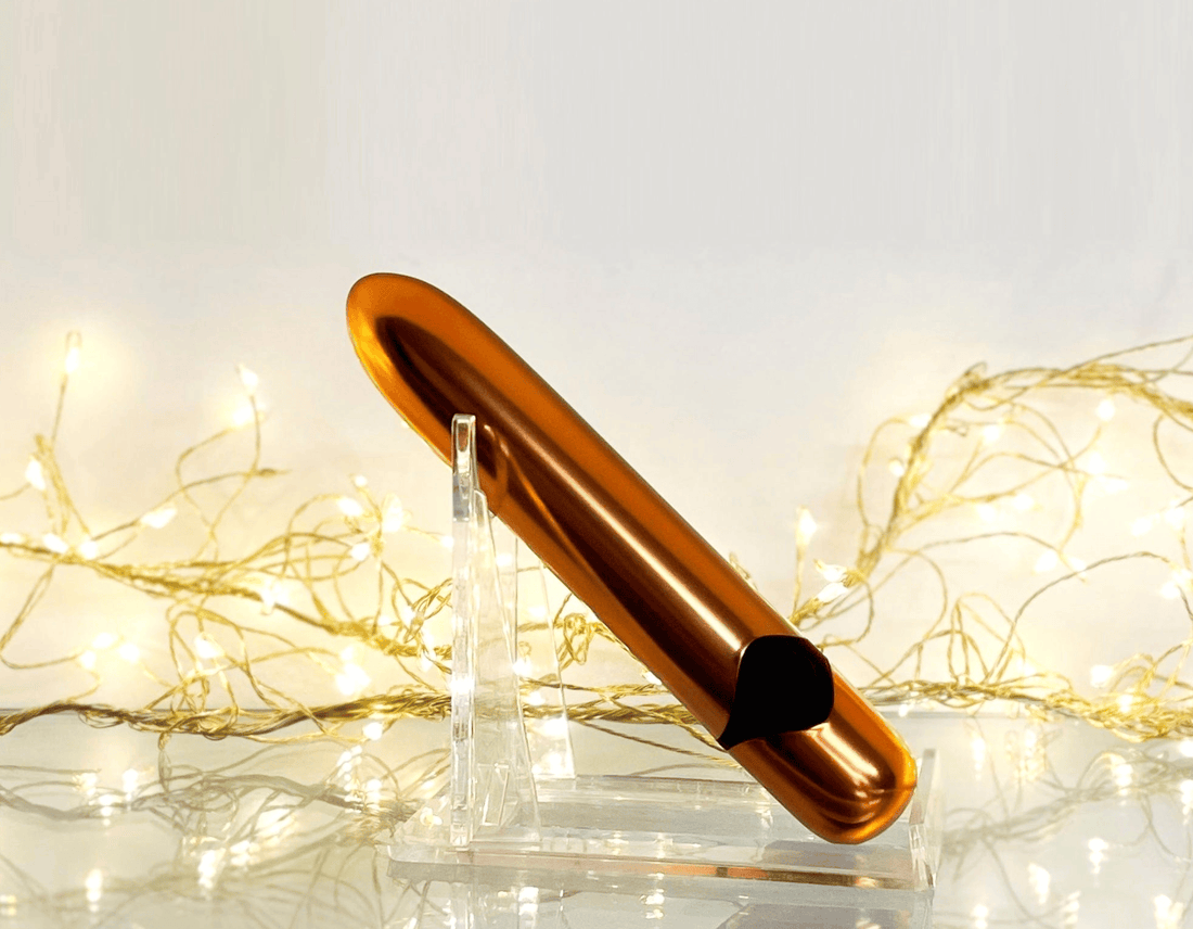 A metallic copper bullet vibrator resting on a clear acrylic stand, set against a warm, softly lit background with glowing fairy lights.