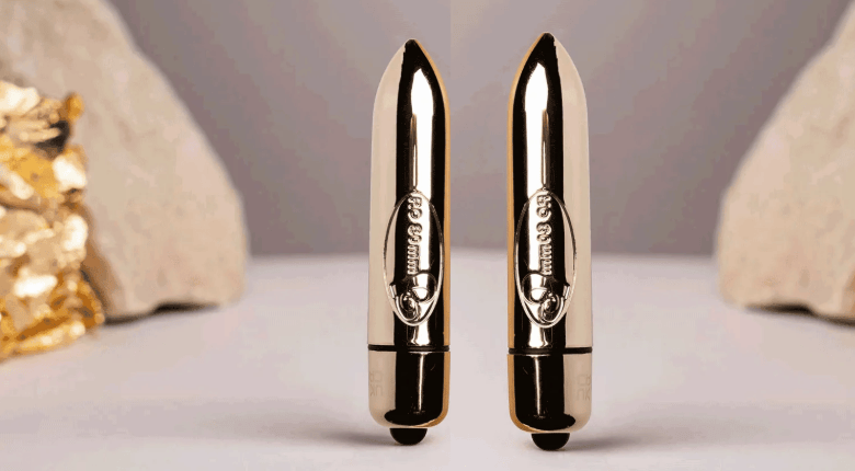 Two metallic gold Rocks Off Ro-80 bullet-vibrators standing upright on a light surface, with a soft gray background and decorative stones on either side.