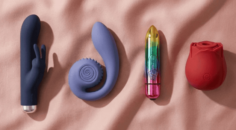 Four intimate wellness vibrators arranged on a soft pink fabric background: a dark navy rabbit vibrator,, a lavender spiral-shaped vibrator, a rainbow bullet, and a red rose suction toy.