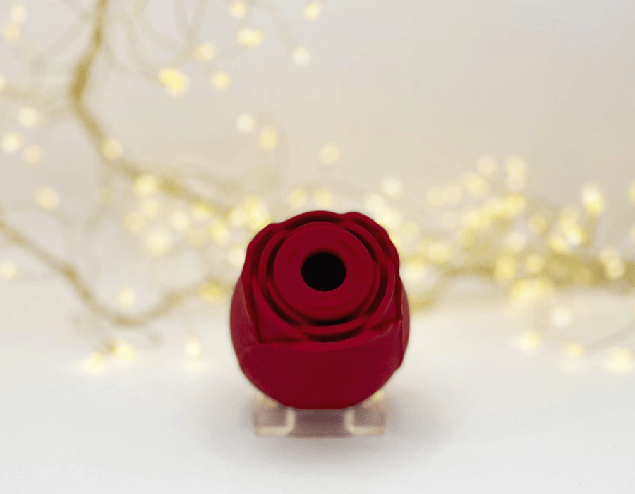A rose clitoral suction toy displayed on a small clear acrylic stand, set against a warm, softly blurred background with glowing fairy lights.