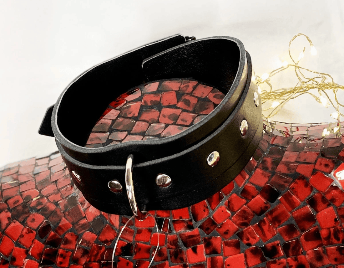 A black leather collar with silver metal studs and a silver metal ring, displayed on a red mosaic background.