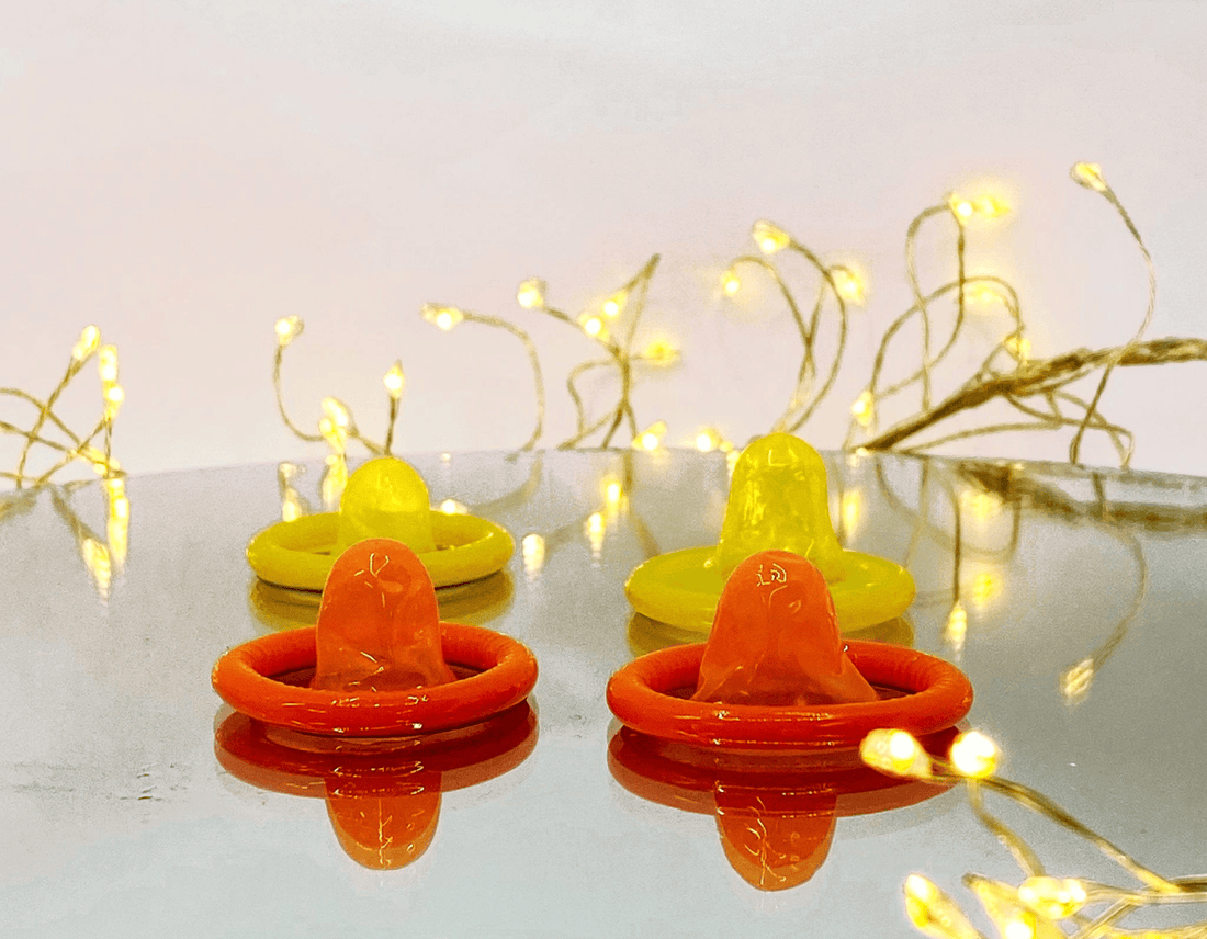 Two yellow condoms and two orange condoms, arranged on a reflective surface with warm fairy lights in the background.