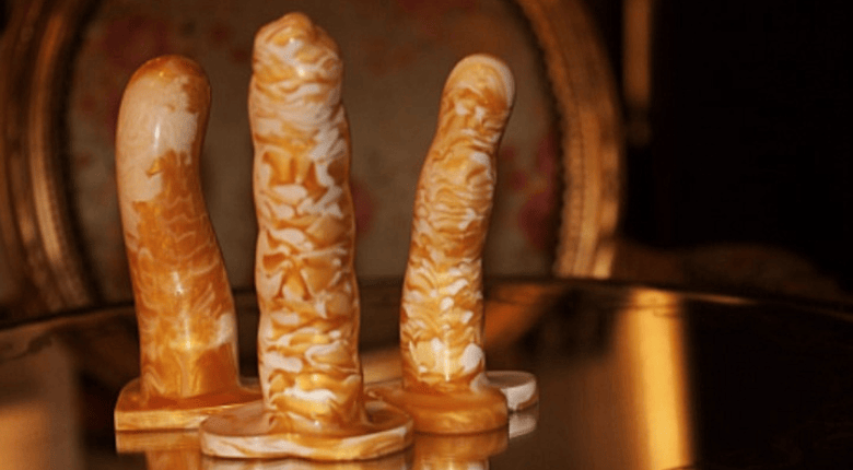 Three Sh! unique silicone dildos in marbled gold, displayed on a reflective dark surface with a blurred ornate background.