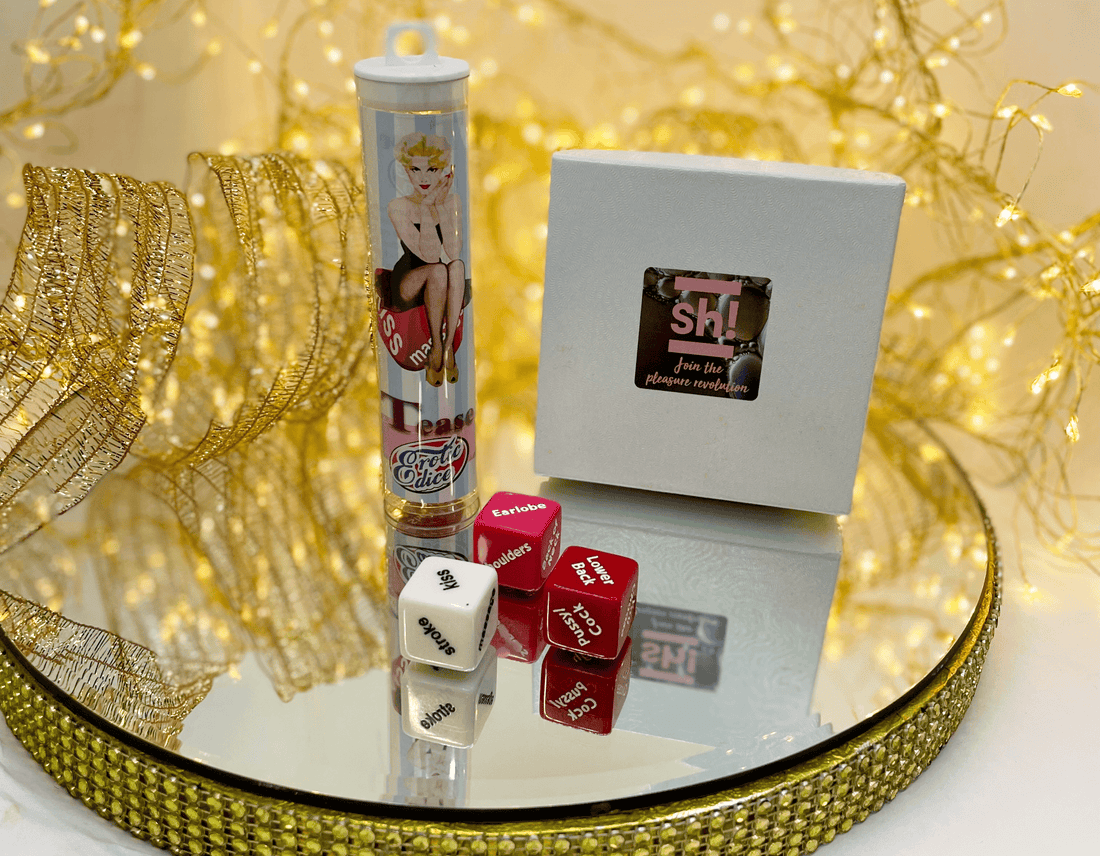 A display of adult gigts & games on a mirrored tray with gold decorative mesh and string lights. The set includes a white Sh! gift Box, Tease erotic dice with' kiss' 'earlobe' &' lower back'