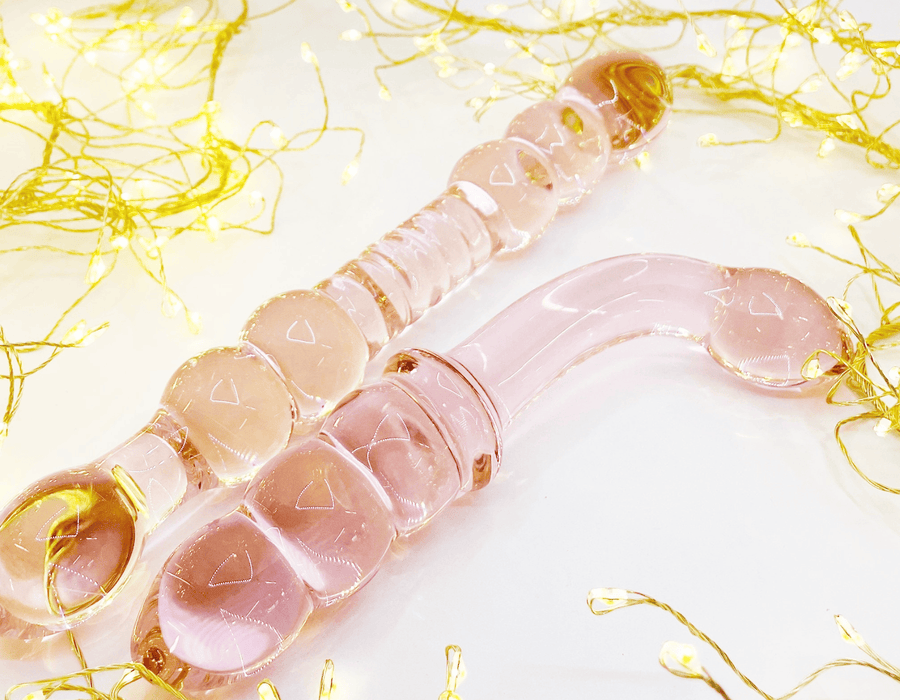 A pair of translucent pink glass dildos, displayed on a white surface with decorative gold fairy lights.