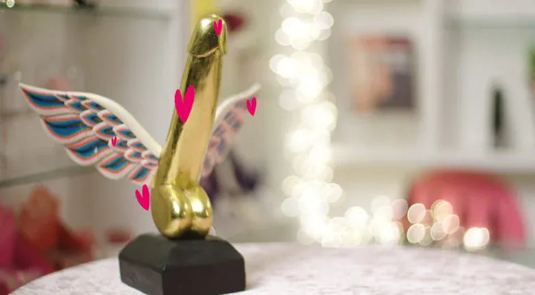 A gold male-penis-shaped trophy with blue and pink wing details, mounted on a black base and displayed on a table in a softly blurred indoor setting.