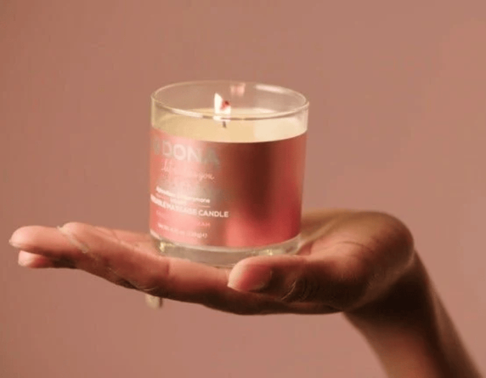 A lit glass jar massage candle held in an open hand against a solid dusty pink background.