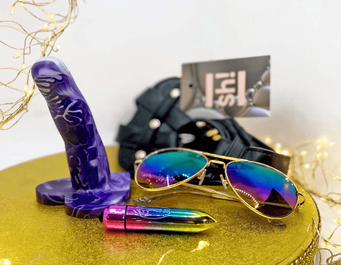 A selection of lesbian sex toys - a purple marbled dildo, a rainbow  bullet vibrator and a black harness, arranged with rainbow aviator sunglasses, on a gold glittery round tray with decorative fairy lights.