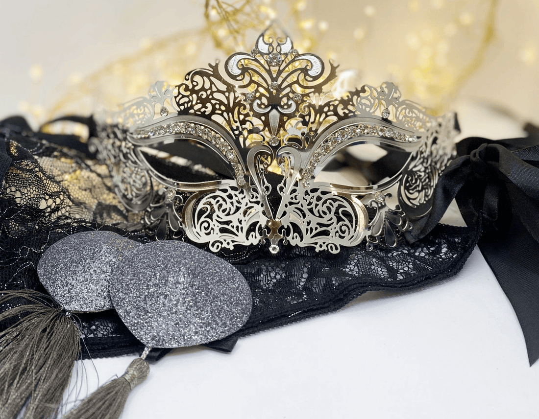 An ornate silver masquerade mask with intricate filigree detailing, displayed on  black lace panties with black ribbons and gray glitter nipple pasties