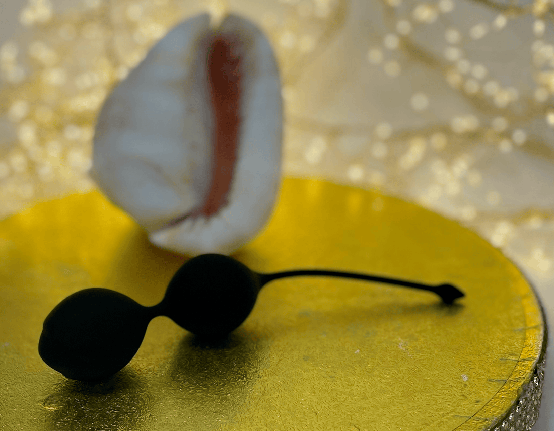 Black silicone kegal ball intimate toy on a gold textured circular tray with a white seashell and warm bokeh lights in the background.