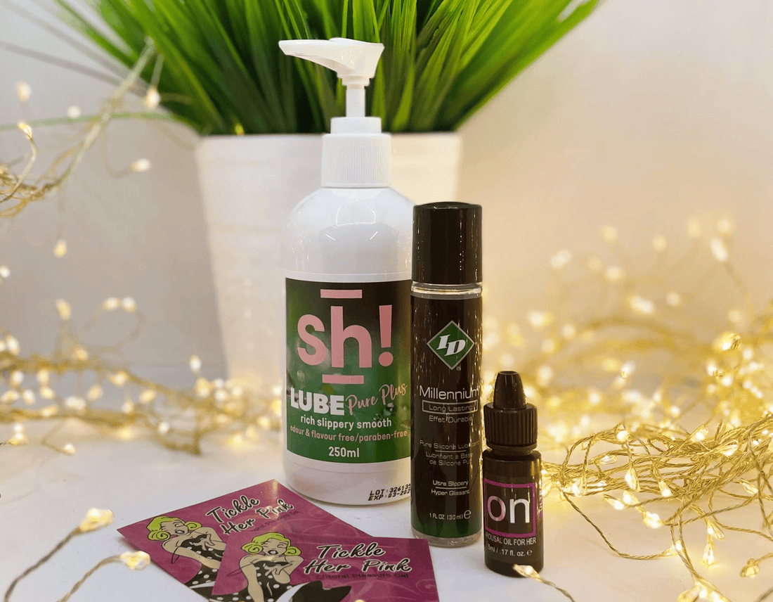 Three personal lubrication products arranged on a white surface with decorative fairy lights and a potted plant in the background: a large white pump bottle labeled 'sh! LUBE', a tall dark bottle labeled 'Millenium', and a small dark dropper bottle labeled 'on'.