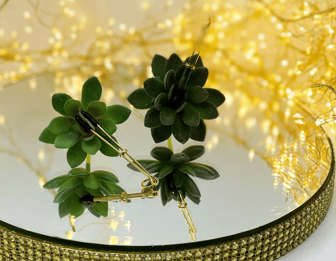 A pair of tweezer nipple clamps attached to a succulent plant, arranged on a round mirrored tray with a gold beaded rim, set against a warm background of glowing yellow fairy lights.