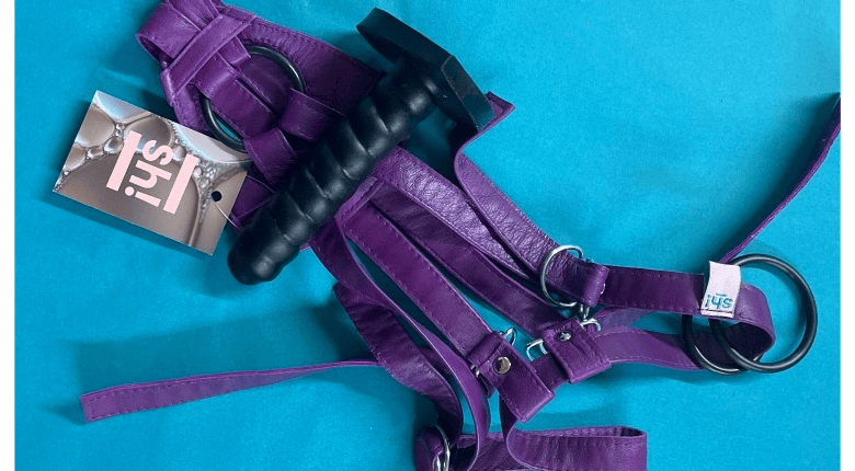 A purple leater 2-strap Sh! Harness with black Wirly 2 silicone dildo on bright blue background