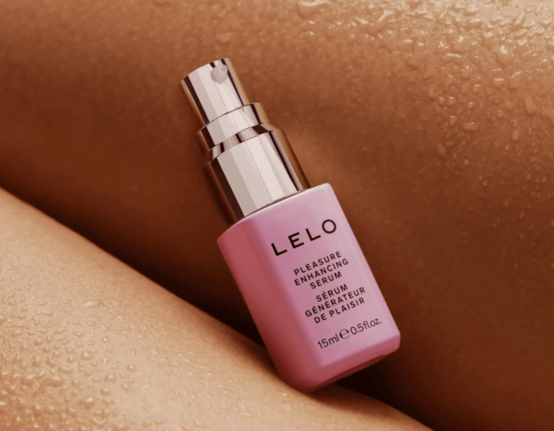 A pink bottle of LELO Pleasure Enhancing Serum resting on warm-toned skin with water droplets.