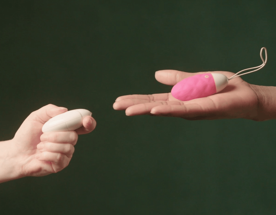 One holding small  pink vibrating egg with another hand  pressing a button on a white remote-control.