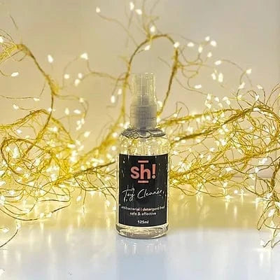 Bottle of  Sh! Sex Toy Cleaner with decorative lights in the background