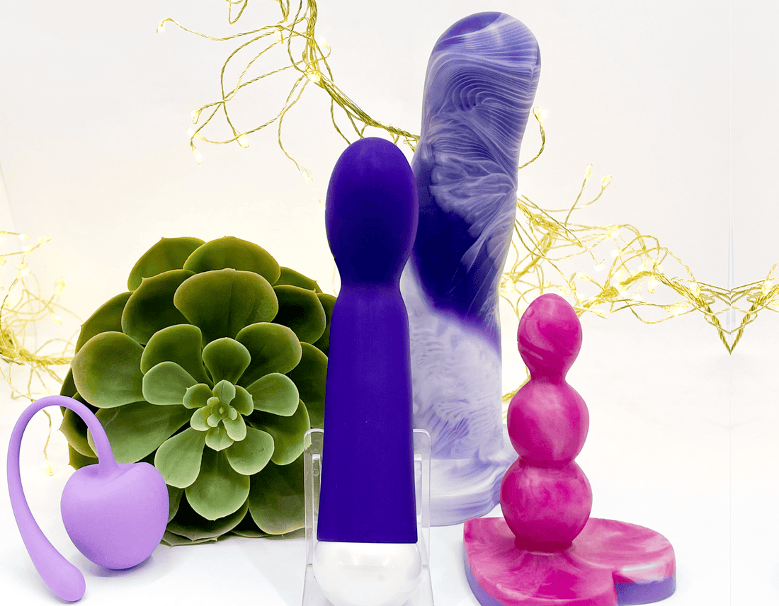 Four different sex toys displayed on clear stands against a white background with decorative gold fairy lights and a green succulent plant. The products include a purple silicone kegal ball, a purple-and-white marbled  purple curved dildo, a pure purple g-spot vibrator and a pink-and-white marbled beaded butt plug.