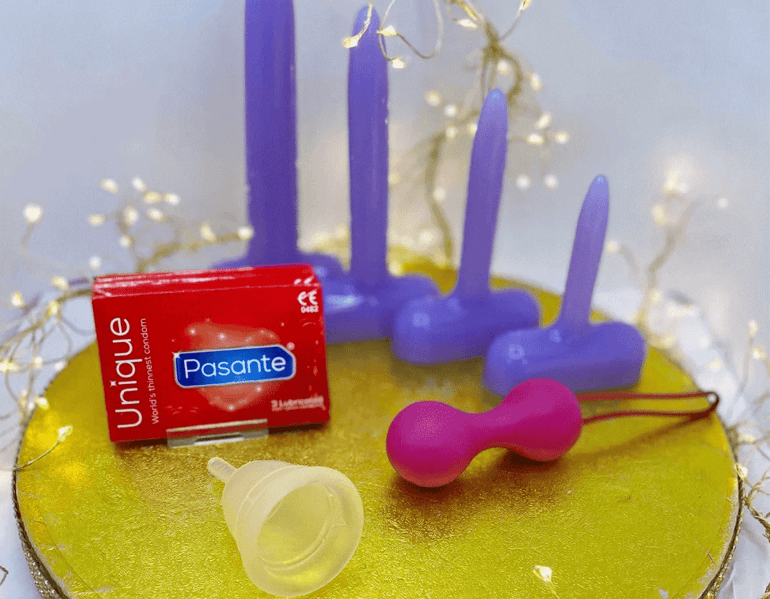 A set of intimate wellness products arranged on a round gold tray, including a red box labeled Unique Pasante, a clear mentrual cup, a pink kegal balls  and purple silicone dilators, with warm fairy lights in the background.