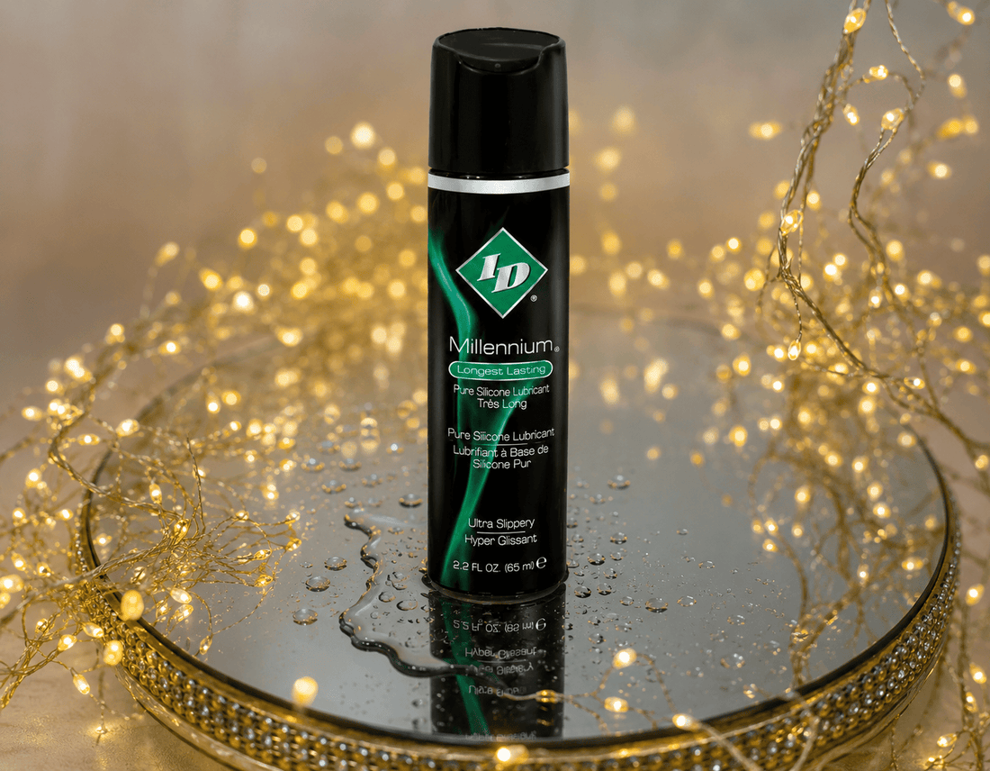 A black bottle of ID Millennium Premium Silicone Lubricant stands on a reflective mirrored tray with gold fairy lights and warm bokeh in the background.