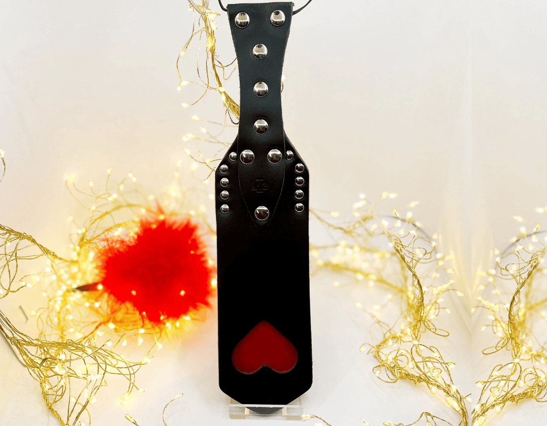 A black leather spanking paddle tawse with silver metal studs and a red heart-shaped cutout, displayed against a white background with decorative gold fairy lights and a red pom-pom.