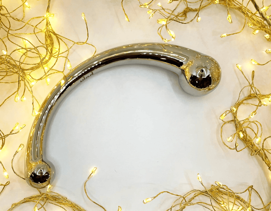 A polished stainless steel double dildo, displayed on a white surface surrounded by glowing warm yellow fairy lights.