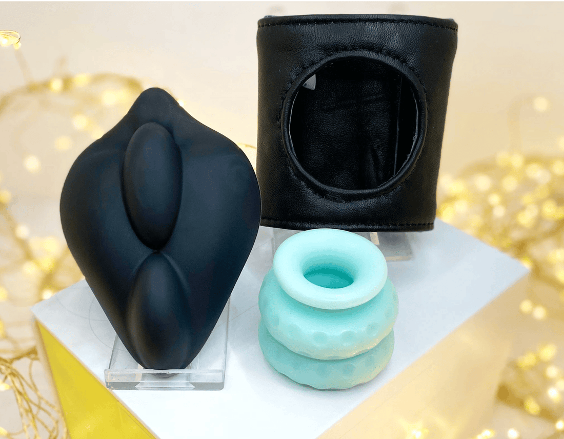 Three israp-on accessories on clear acrylic stands: a black silicone bananapants dildo base, a dual-dildo cuff to add an extra dildo during strap-on play and an oh-nut ring.