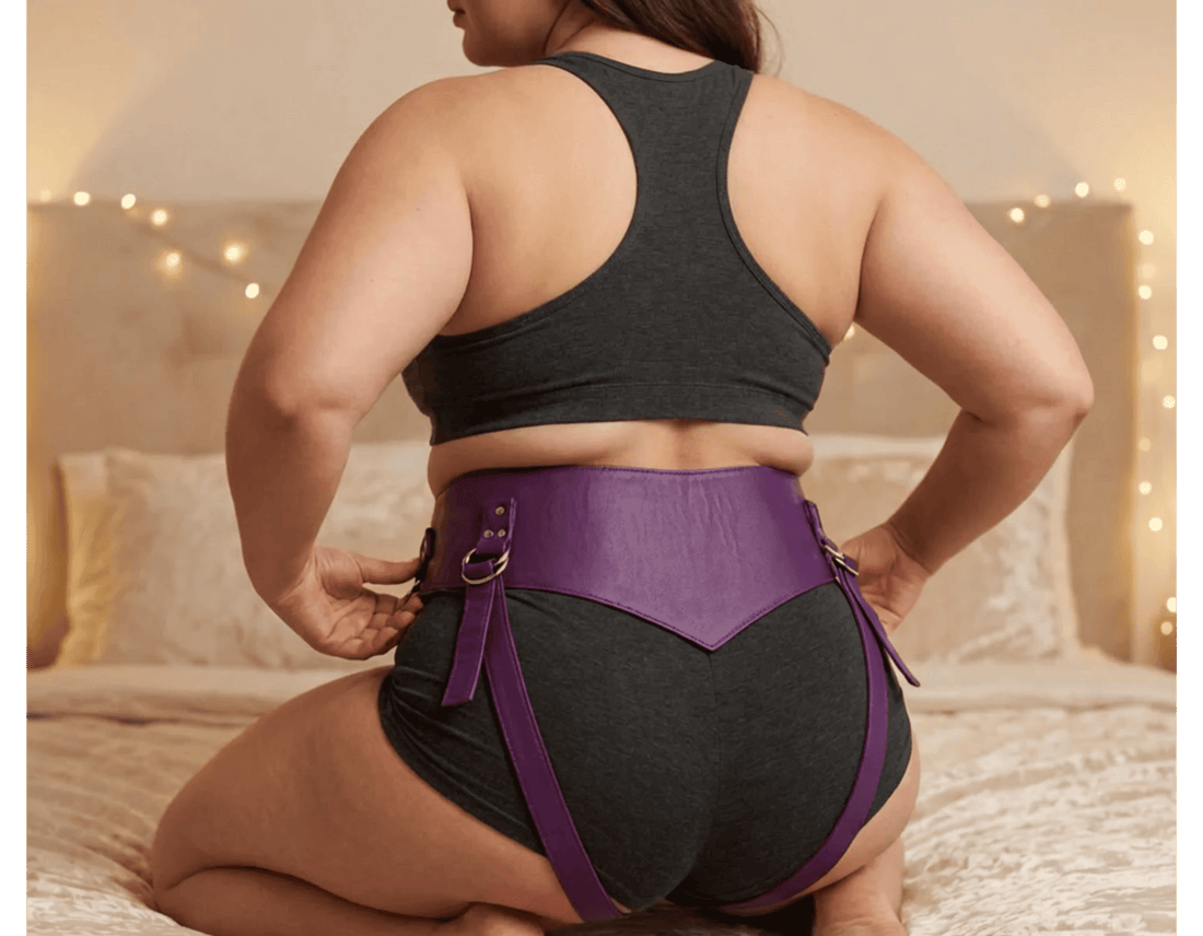 A woman kneels on a bed showing the back of a purple leather super strapon harness by Sh! Women's Store.