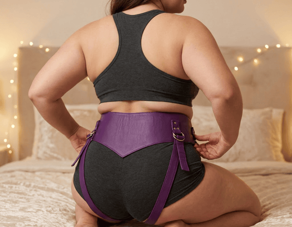 A  woman kneeling on a bed, viewed from behind, wearing a dark gray sports bra and high-waisted shorts with a purple leather strap-on harness.