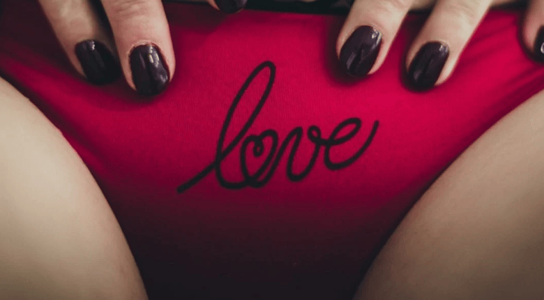 A close-up of a person's hands with dark brown manicured nails resting on bright pink underwear featuring the word 'love' printed in black script across the front.
