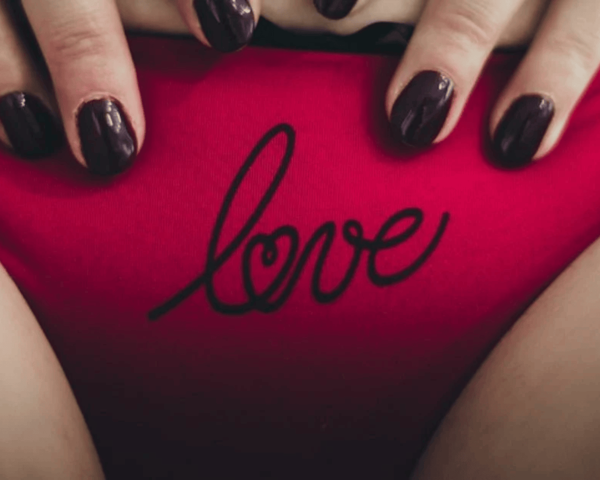 A close-up of a person's hands with dark plum nail polish resting on bright pink underwear featuring a black cursive 'love' graphic.