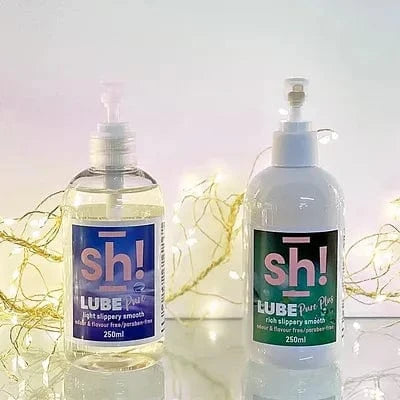 Two bottles of 'sh!' Lube on a reflective surface with a soft, blurred background.