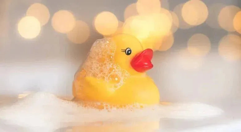 A yellow rubber duck with a red beak sitting in white foam, with a warm bokeh background of soft golden lights.
